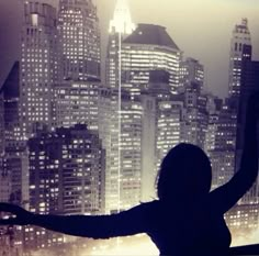 a woman standing in front of a window with her arms spread out and the city lights behind her