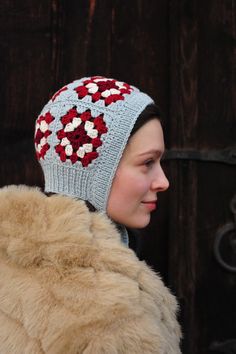 a woman wearing a knitted hat with red and white flowers on the top, standing in front of a wooden door