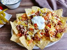 a white plate topped with nachos covered in cheese and toppings next to a bowl of sour cream