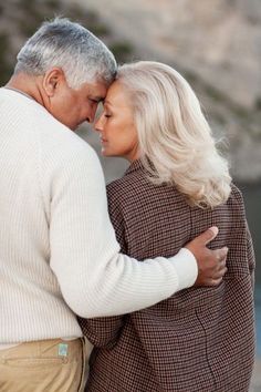 an older couple embracing each other in front of a body of water with rocks behind them