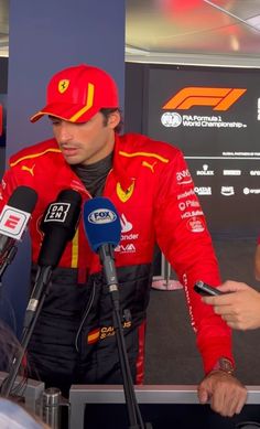 a man in a red racing suit talking to the media with microphones around him