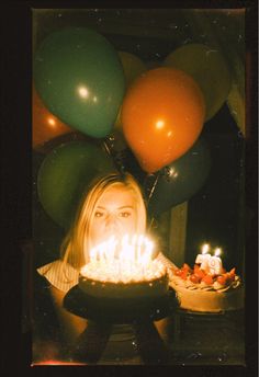 a woman holding a cake with lit candles in front of her face, surrounded by balloons