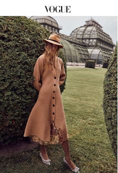 a woman in a brown coat and hat standing next to some bushes with greenhouses in the background