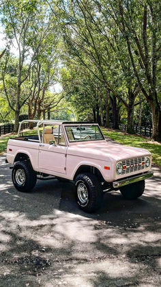 A classic Ford Bronco in a unique Rosé pink color, parked on a shaded road lined with lush green trees. The Bronco features a white soft top, large off-road tires, and a clean, well-maintained body, showcasing its retro charm and stylish color palette. Classic Car Convertible, Pink Ford Truck, Cute First Cars, Dream Car Jeep, Vintage Ford Bronco, Old Bronco, Bow Art