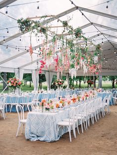 an outdoor tent with tables and chairs covered in blue linens, flowers and greenery hanging from the ceiling