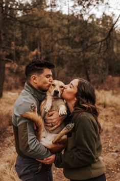 a man and woman are kissing their dog in the woods while they hold it up