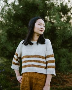 a woman standing in front of some trees wearing a white and brown sweater with stripes on it