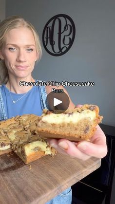 a woman holding a piece of cake on top of a wooden cutting board with chocolate chip cheesecake