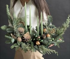 a woman holding a christmas wreath with pine cones and evergreens on it, surrounded by candles