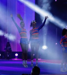 two cheerleaders are performing on stage with their arms in the air and lights behind them