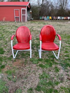 two red chairs sitting on top of a grass covered field in front of a barn