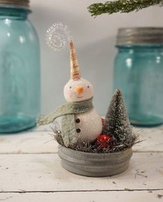 a snowman sitting in a bowl next to some glass jars and a pine tree