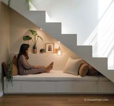 a woman sitting on a white couch under a stair case next to a potted plant