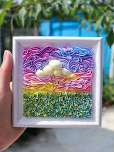 a hand holding up a small paper art piece with swirls on it and clouds in the background