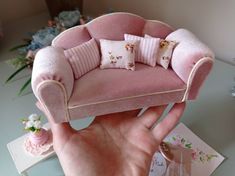 a hand holding a miniature pink chair with pillows on it's back and two small flowers in the background