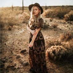 a woman in a long dress and hat standing in the desert with her arms crossed