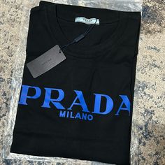 Prada Milano Black T-Shirt Blue Lettering Black Designer Logo Crew Neck Top, Black Crew Neck Top With Designer Logo, Black Crew Neck Tops With Designer Logo, Black Designer Logo Top For Streetwear, Black Cotton Tops With Designer Logo, Designer Blue Crew Neck T-shirt, Designer Blue Tops With Logo Print, Designer Blue Tops For Streetwear, Designer Blue Cotton T-shirt