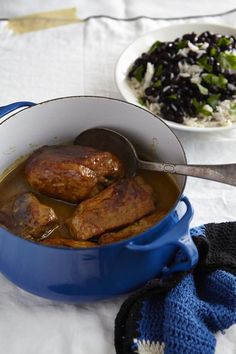 a blue pot filled with meat and beans next to a bowl of rice on a table