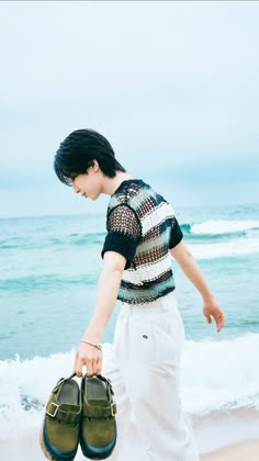 a young man holding two pairs of shoes on the sand at the ocean's edge