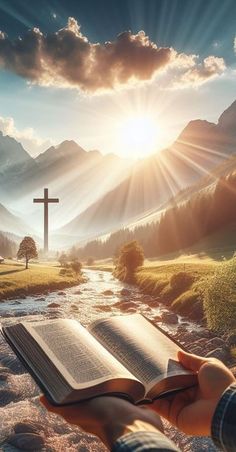 a person holding an open book in front of a cross on a mountain with a stream running through it