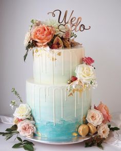 a three tiered cake with flowers and gold lettering on the top, sitting on a table