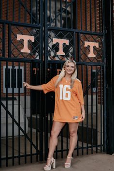 a woman standing in front of a gate wearing an orange football jersey and heels with her arms out