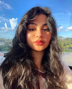 a woman with long dark hair and orange lipstick looks into the camera while sitting in a car