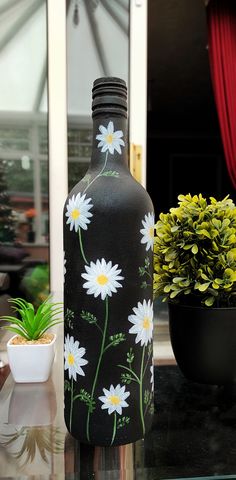 a black vase with white daisies painted on it sitting next to a potted plant