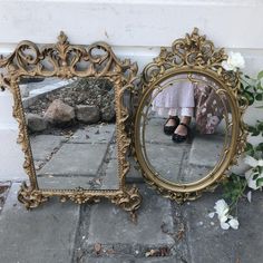 a woman's reflection in an ornate gold framed mirror on the ground next to flowers