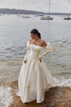 a woman standing on top of a rock near the ocean wearing a white dress with long sleeves