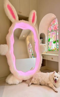 a white dog laying on the floor in front of a mirror with pink bunny ears