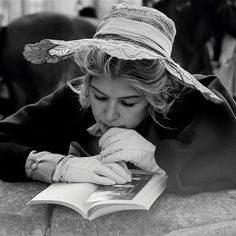 black and white photograph of a woman wearing a hat reading a book while laying on the ground