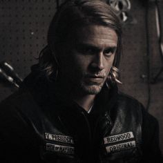 a man with long hair wearing a leather jacket and looking at the camera while standing in a garage