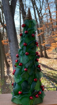 a green christmas tree with red berries and pine cone on top sitting on a wooden table in front of some trees