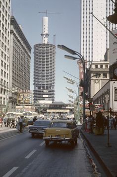 an old car driving down the street in front of some tall buildings and people walking on the sidewalk