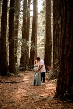 a man and woman standing in the middle of a forest with tall trees behind them