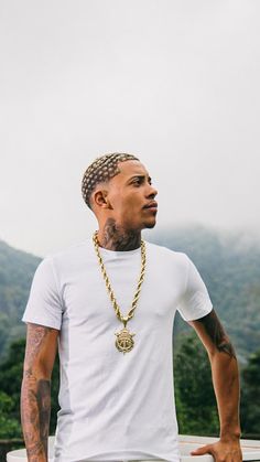 a man wearing a white t - shirt and gold chains standing in front of mountains