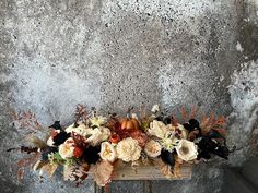 an arrangement of flowers on a table against a concrete wall with leaves and pumpkins