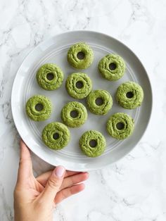 a white plate topped with green donuts on top of a marble countertop next to a person's hand