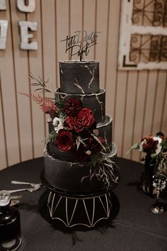 a three tiered black cake with red flowers and greenery sits on a table