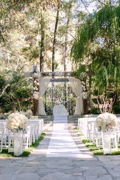 an outdoor ceremony setup with white chairs and flowers on the aisle, surrounded by trees