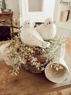 two white birds sitting on top of a vase filled with flowers and greenery in front of a fireplace