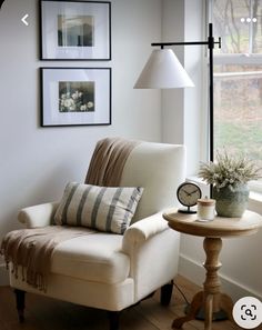 a white chair sitting next to a table with a lamp on it and pictures above