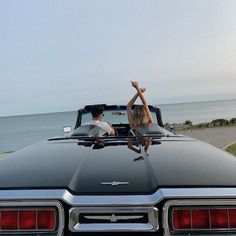 two people sitting in the back of a convertible car with their hands up and one person standing on the roof