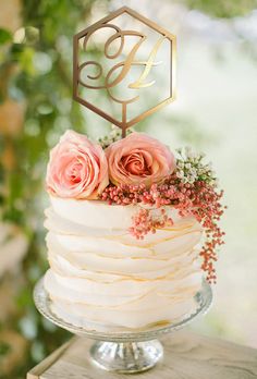 a white cake with pink flowers on top and a gold monogram decoration above it