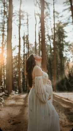 a pregnant woman in a white dress standing on a dirt road surrounded by tall trees