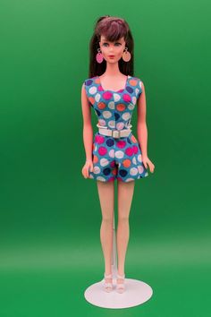 a doll is standing on a green background wearing a polka dot dress and pink earrings