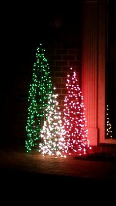 two christmas trees are lit up in front of a brick building at night with red and green lights