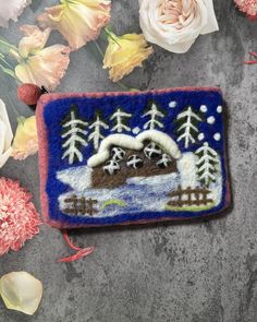 an embroidered brooch with trees and snow in the background next to flowers on a gray surface