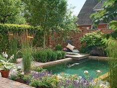 a garden with a pond surrounded by trees and flowers on the side of a house